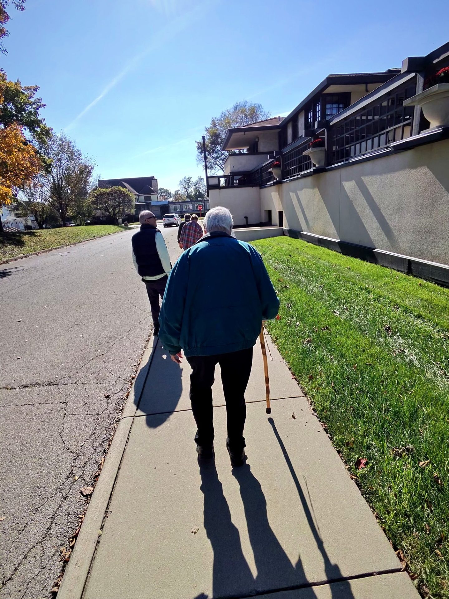 Elderly man with white hair and cane walking along sidewalk beside Westcott House Prairie wall with red flower planters