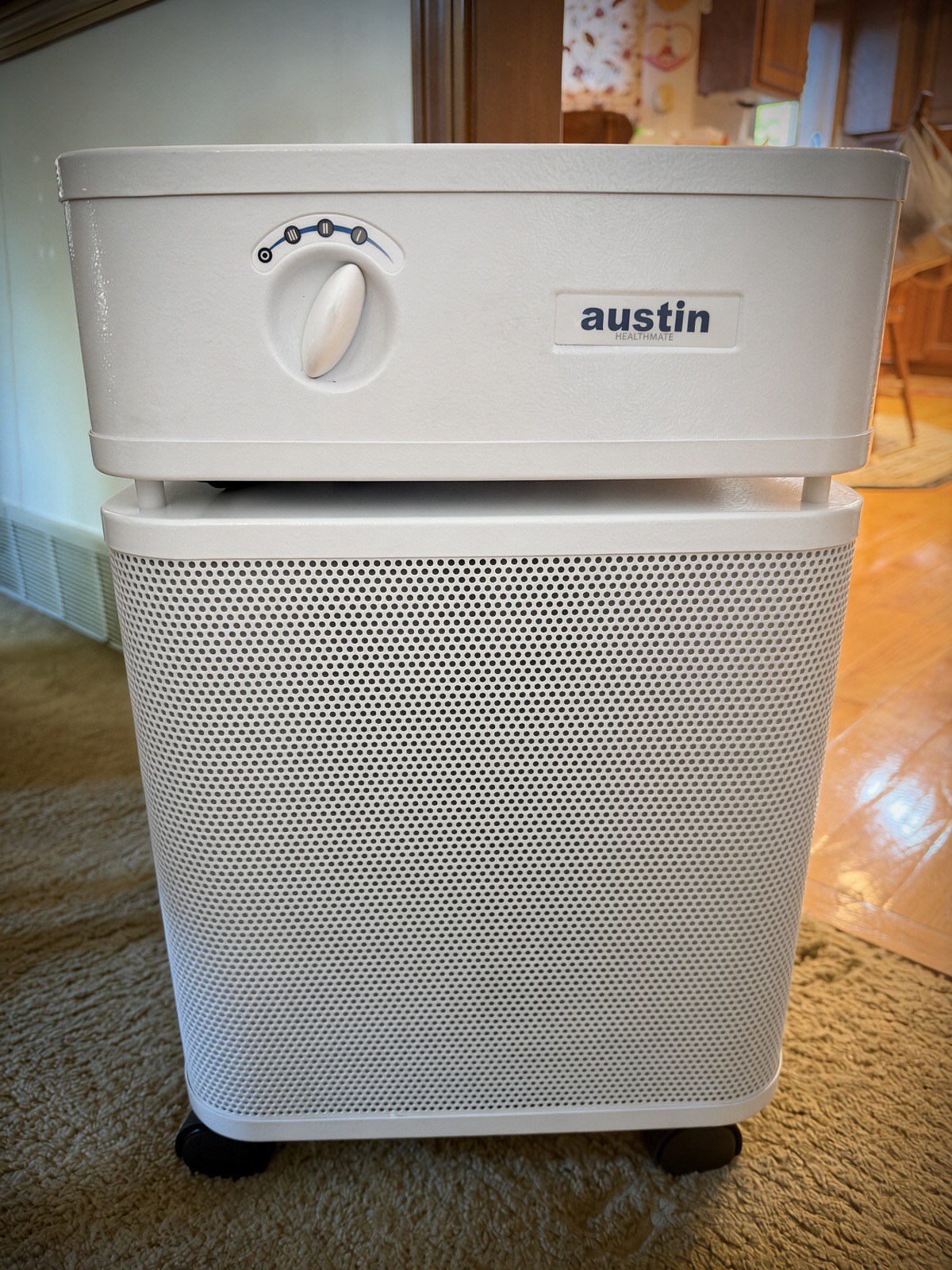 Front view of a white Austin Air HealthMate air purifier with its dial control and austin logo, on casters on a carpet next to a hardwood floor