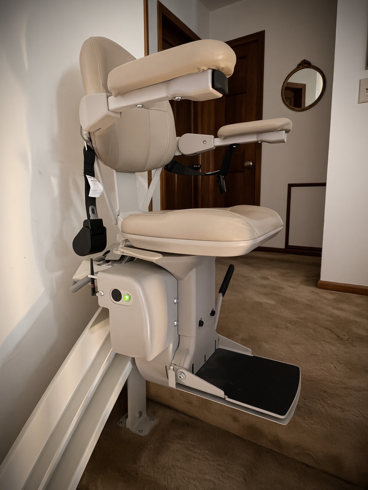 Beige stair chairlift with padded seat, armrests and seat belt parked at the top of a carpeted staircase