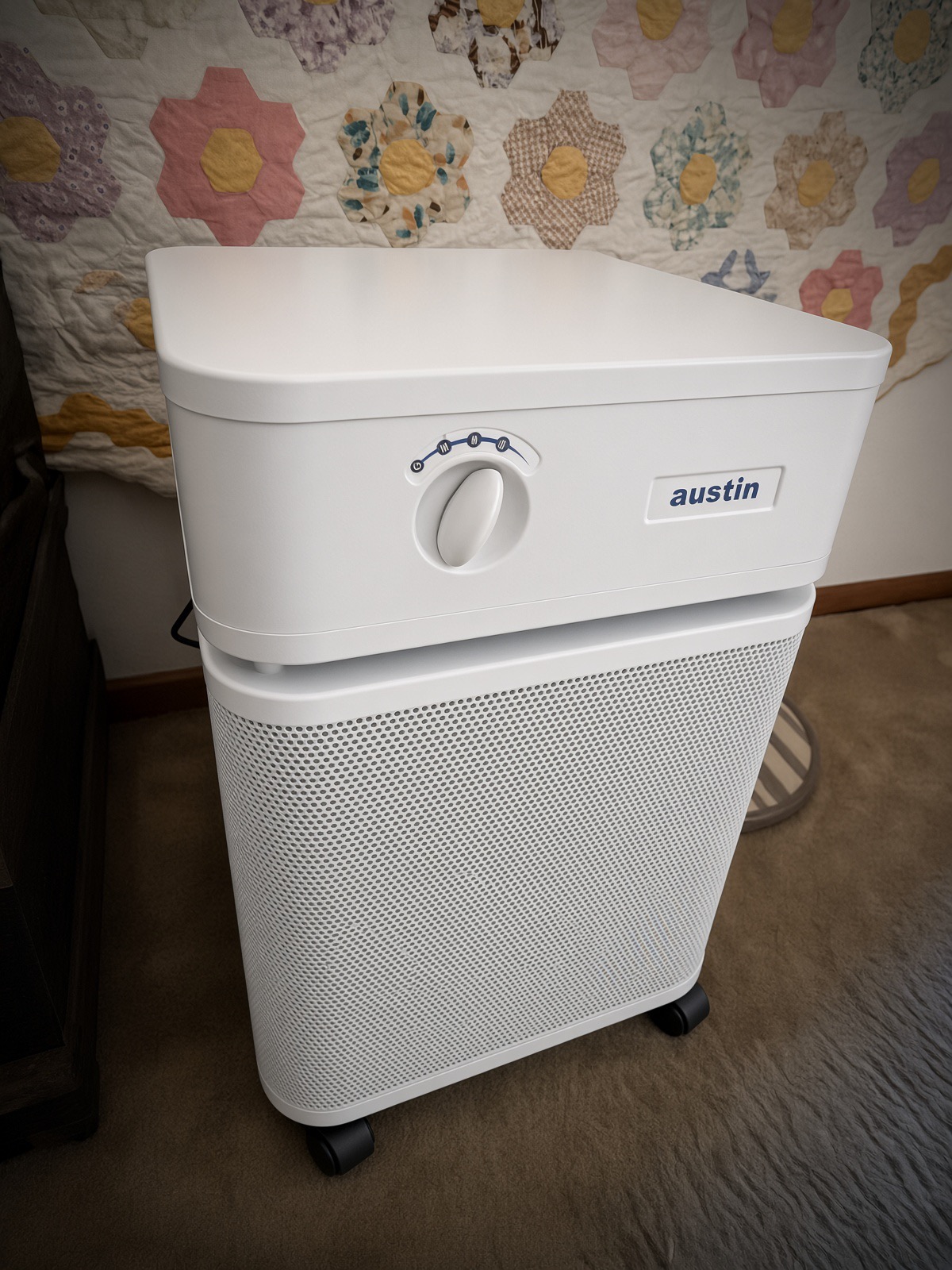 White Austin Air HealthMate air purifier on casters on carpet in front of a flower garden patchwork quilt hanging on the wall