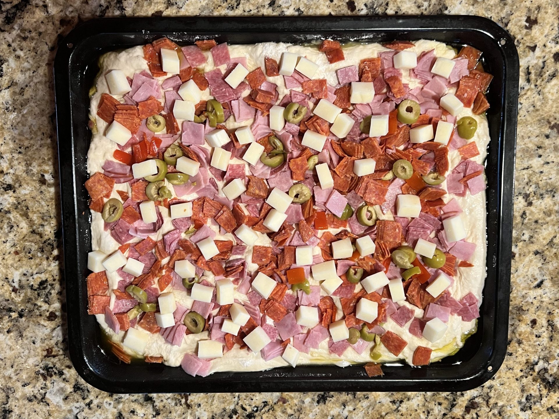 Focaccia dough in a pan topped with Italian meats, cheese and green olives, before baking