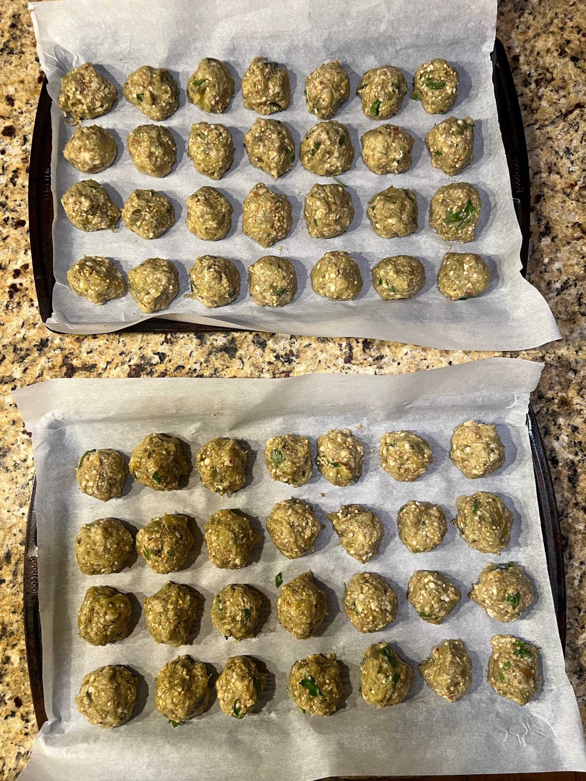 Two parchment-lined sheet pans of raw rolled Italian eggplant balls, ready for the oven
