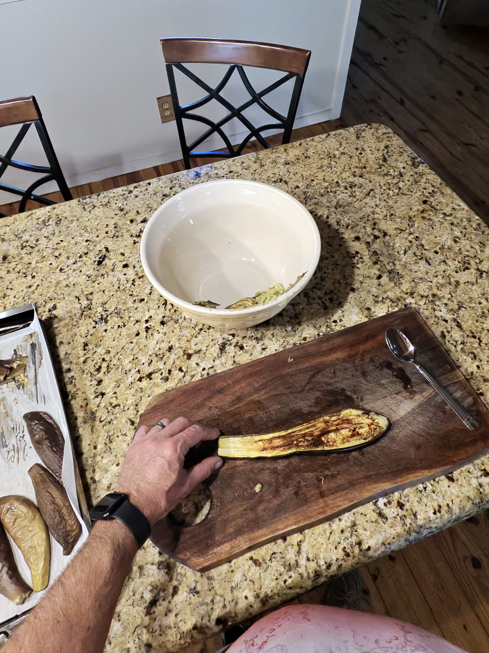 Scooping roasted eggplant flesh out of the skin with a spoon on a wooden cutting board